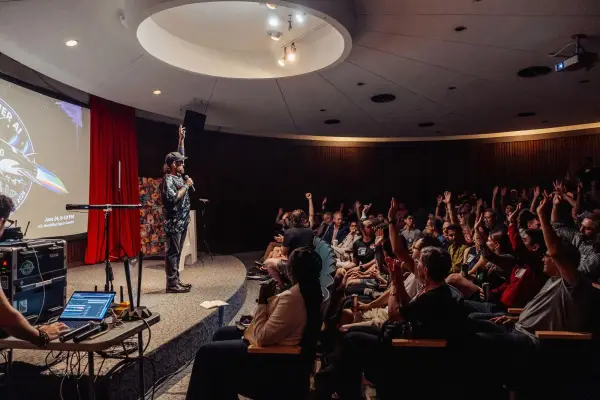 Kris Krüg raises one hand onstage while rows of attendees raise their hands at the H.R. MacMillan Space Centre