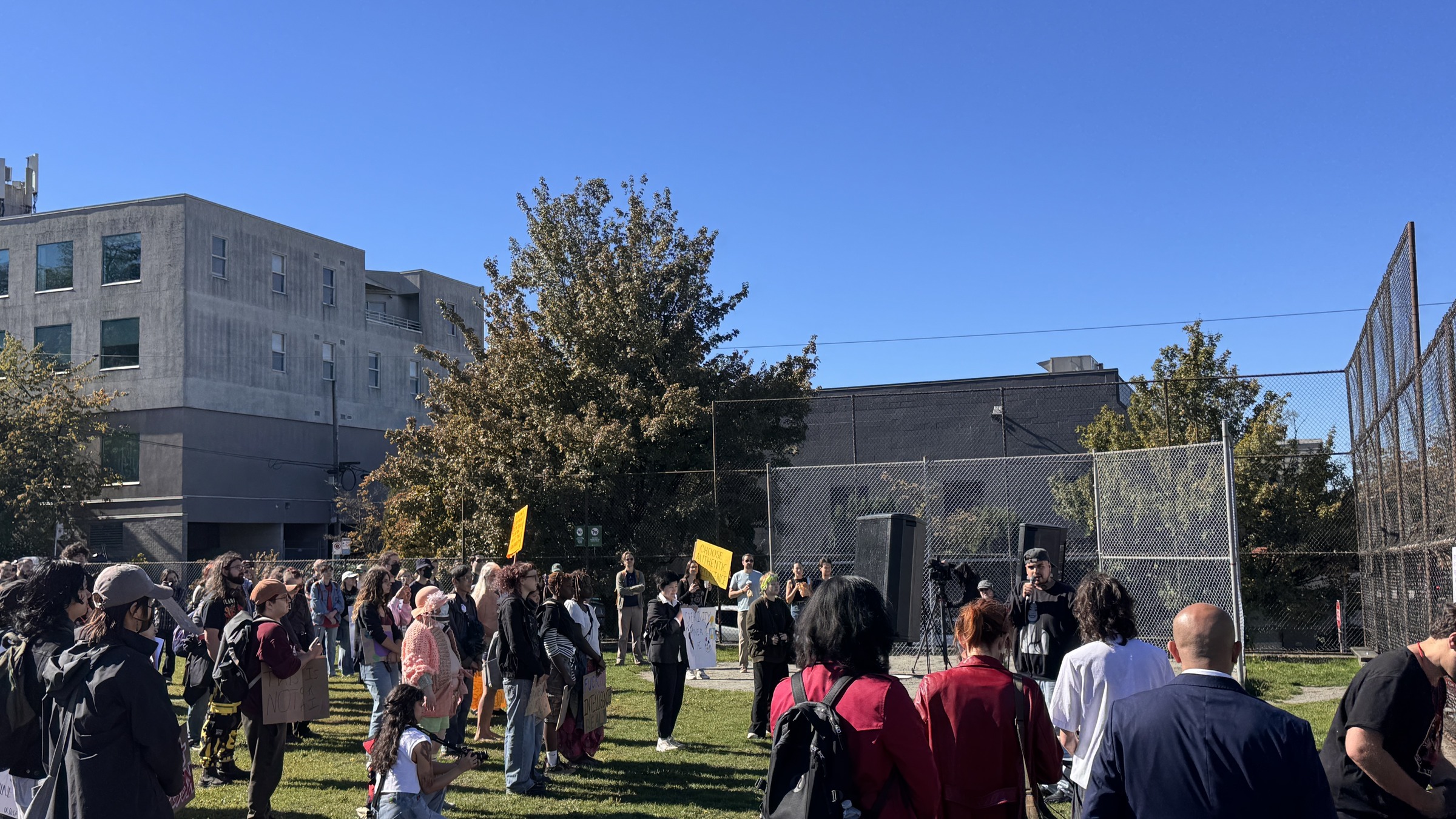 People gather around a speaker beside the fence, with handmade placards held among the crowd.